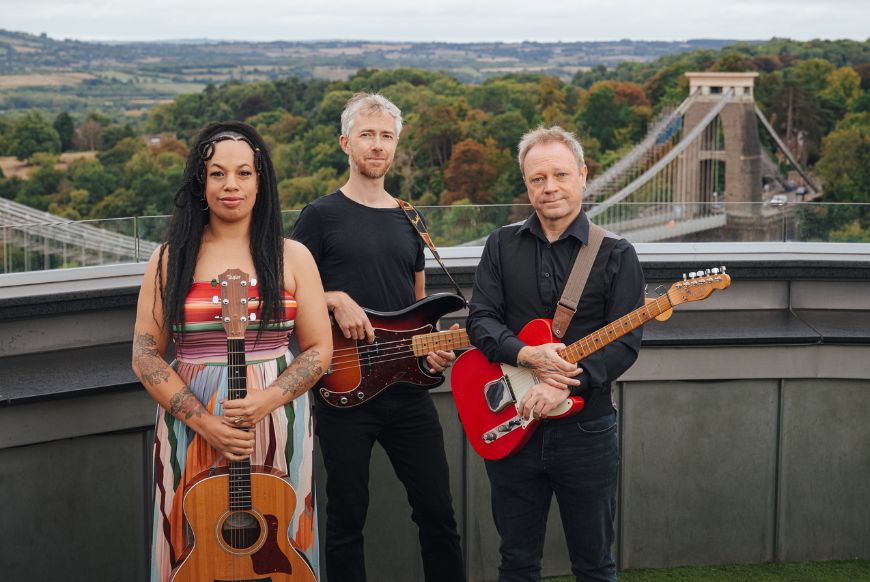 three people, one woman in a brightly coloured dress and two men in all black. Stand overlooking the bristol suspension bridge while holding guitars.